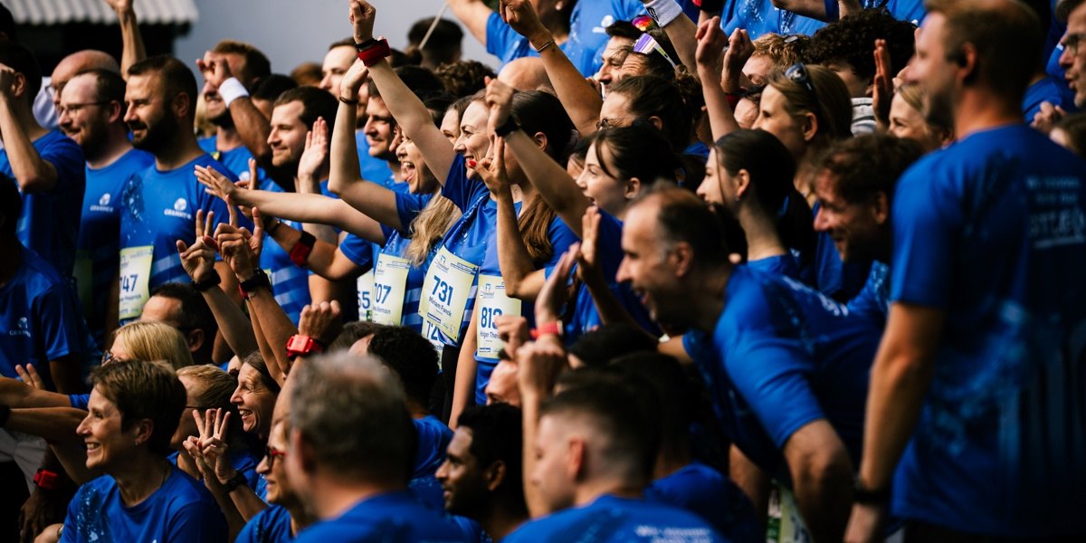 Teilnahme jetzt planen Eine große Gruppe lachender Läufer:innen in blauen Shirts mit Startnummern jubelt gemeinsam mit erhobenen Armen in die Kamera.